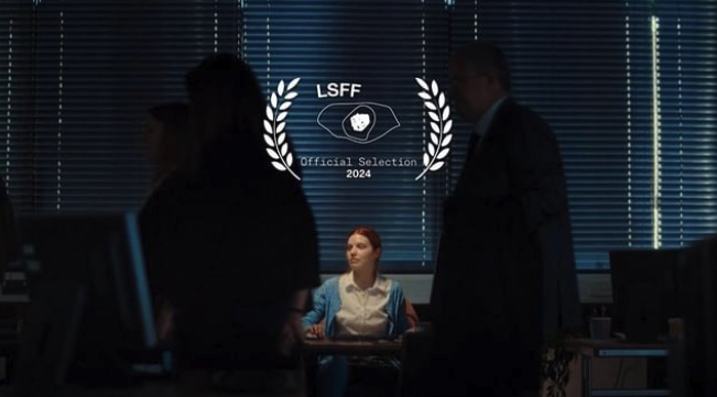 A dark shot of an office with 2 people overlooking an office worker. Lots of film grain. A logo is overlayed which says London Short Film Festival 2024.